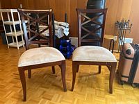 Two matching wooden dining chairs with beige cushioned seats and X-back design, placed side by side on parquet floor.