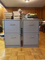 Two grey steel filing cabinets side by side with several books stacked on top of them, placed indoors on parquet flooring against a wood panel wall.