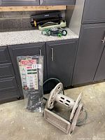 Overall view of the garden hose reel, nozzles, sprinklers on countertop, and plastic garden obelisk in packaging leaning on cabinet.