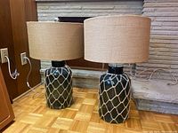 Two matching tabletop lamps with dark glass bases covered by a tan rope netting, beige drum shades on top, placed on a parquet floor near a stone fireplace.