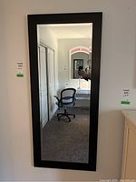 Frontal view of full-length rectangular mirror with black wooden frame hung on a wall, reflecting a desk and chair.