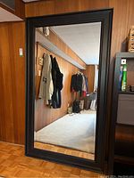 Full front view of the full length mirror leaning against a wall, showing its size, black wood frame and beveled edges.