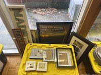 Various picture and poster frames and decorative signs arranged on and behind yellow plastic tote bin near window, showing size and style variety.