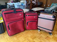 Three luggage pieces on parquet floor including two red soft-sided suitcases and one rose gold London Fog roller bag.
