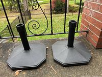 Pair of black outdoor umbrella stands shown side by side on a patio surface with iron railing and green lawn in background.