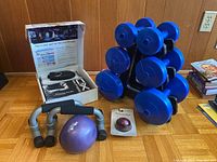 Fitness kit with P90X home fitness guide box open showing DVDs and accessories, plus push-up handles, small purple exercise ball and massage roller on hardwood floor