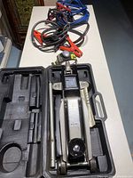 Photo showing hydraulic floor jack inside black plastic carrying case and two pairs of heavy duty jumper cables with clamps on white table.