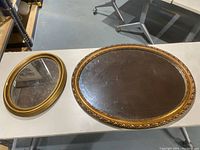 Two oval gold framed mirrors side by side on a table, large mirror approx 30 inches and smaller one approx 18 inches with visible chip on frame