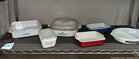 Full shelf showing all vintage Corningware and Pyrex cookware including covered and uncovered casseroles, pink Gooseberry refrigerator dish, and divided vegetable dish needing cleaning.