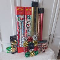 Overview photo showing all Christmas gift wrap rolls in various themes including kids, gold color, and luxury red craft wrap. Large packs of bows in assorted colors and various sized ribbons visible, along with boxes of themed gift tags and Scotch tape.