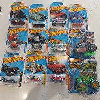 Full lot showing all 12 cars and 2 monster trucks in sealed packaging on white surface