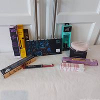 Full lot of 10 makeup products displayed on a white surface with closet doors behind. Products include Urban Decay setting spray, Got2b brow gel, Lancome mascara, Uoma eyeshadow palette, e.l.f. primer, Laura Mercier loose powder with puff, Winky Lux brow pencil, Kylie Jenner lip gloss, Revlon lip crayon.
