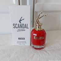 Photo showing the red and clear faceted perfume bottle with gold-tone legs-shaped cap next to the plain white tester box with black branding text.