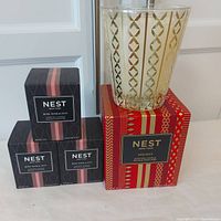 Three boxed Nest Fragrances Rose Noir & Oud scented candles and one large Nest Fragrances Holiday scented candle in decorative glass on a white table.