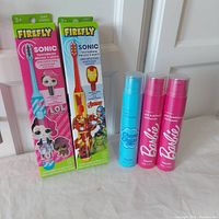 Two Sonic Firefly battery powered sonic toothbrushes in original packaging with antibacterial covers, one pink LOL Surprise and one Avengers themed, alongside three Hismile fluoride toothpaste tubes: one blue Chupa Chups cola flavored and two pink Barbie bubblegum flavored.