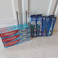 Packaged Oral B Pro 100 Crossaction (black) and Gumcare (white) toothbrushes, two Oral B kids toothbrushes with color changing bristles and six Colgate Max Fresh toothpaste tubes arranged on table.