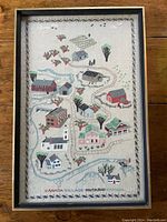 Full view of framed vintage needlepoint sampler featuring a small village map with buildings and natural elements.