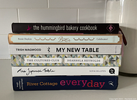Six cookbooks stacked with spine titles visible: The Hummingbird Bakery Cookbook, Butter Celebrates!, My New Table, The Cultured Club, The Japanese Table, River Cottage Everyday.