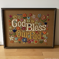 Front view of framed needlepoint featuring 'God Bless Our Pad' text and colorful floral embroidery on tan canvas.