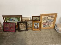 Wide view showing eight framed needlepoint and silk artworks arranged leaning against wall.