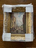 Photo of original signed oil painting on canvas wrapped in protective plastic and corner protectors, showing the gold decorative frame and part of the street scene.
