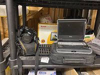 Photo showing Nikon DSLR camera in black and orange carrying case, Kodak EasyShare digital picture frame box, and Philips portable DVD player with carrying case on a shelving unit.