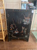 Front view of black lacquer wooden cabinet with detailed paintings showing flowers, trees, birds, pagoda, and Asian figures on double doors with metal ring pulls.