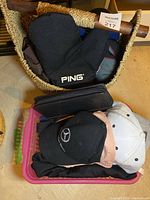 Woven basket containing black Ping glove, black leather wallet, and visible parts of baseball caps on pink plastic tray beneath basket.