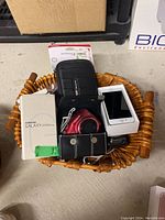 Overview of wicker basket containing various electronics and accessories including Samsung box, Nintendo case, Canon camera, iPhone box, and leather case.