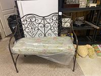 Full view of iron bench showing metal frame with leaf and scrollwork and floral cushioned seat covered in plastic.