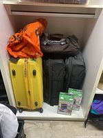 Wide view showing yellow hard-shell suitcase, two black luggage bags, brown fabric travel bag, orange plastic bag, and PAC N Stack storage bags in boxes.