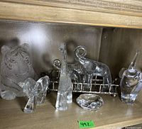 A full shot of the six glass animal figurines displayed on a wooden shelf, showing tiger, giraffe, elephant, rhinoceros, bird, and faceted dish.