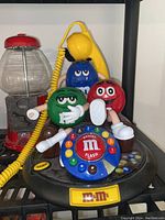 Photo showing the vintage M&M themed rotary dial telephone with yellow handset and coiled cord alongside a red vintage gumball machine.