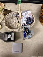 Lot displaying the white wicker basket containing the Mighty Dwarf multimedia speaker box and Acqua Indiglo watch, with Swiss Army watch in case and tin nearby on table.