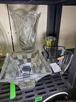 Photo shows large Nachtmann crystal vase at the back left, square glass bowl front left, medium gold trim vase at back right, black rim clear bowl front right on metal shelving.