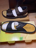 Black and brown Naot sandals shown on top of their respective shoeboxes, with visible stitching detail and cushioned footbeds.