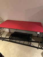 Full view of the folding red table showing red rectangular top and black iron base with mesh shelf underneath on a basement floor.