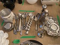 Full view of silver serving items on table including salad tongs, candle holders, pepper mill, cocktail shaker, serving trays, bowls, and nutcracker.