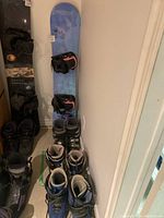 Photo showing side view of snowboard with bindings and three pairs of snowboard boots arranged on floor