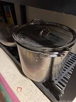 Large stainless steel soup pot with lid and a mixing bowl on a black metal rack, showing size and condition