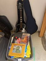 Steel reinforced Stella acoustic guitar inside a soft case against a wall with a plastic tote filled with books in front.
