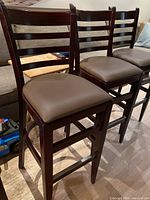Four wooden bar stools with ladder-back design and brown cushioned seats arranged side by side on wood floor.