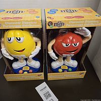 Two M&M Character Dispensers in original packaging side by side, yellow peanut character and red chocolate character visible.
