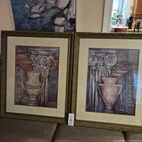 Photo of two large vintage framed prints side by side showing detailed urns and classical architectural motifs in ornate greenish-gold frames with ivory mats.