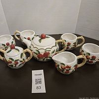 Full view of the tea set showing teapot, sugar bowl with lid, creamer, and four mugs with embossed red apple design and handles resembling tree stems.