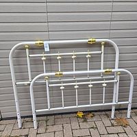 Full view of the vintage white metal and brass bed frame headboard and footboard resting against a siding wall on a paved surface, showing overall shape and condition.