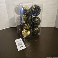 Clear plastic box containing 13 large black ball Christmas ornaments decorated with gold patterns and glitter, approximately 3 inches each.