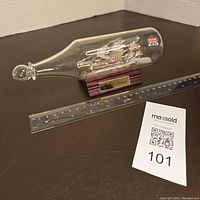 Glass bottle containing detailed glass ship model labeled H.M.S Discovery, placed next to a ruler and auction info card for scale.