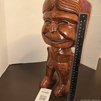 Front view of the heavy hand carved wooden smiling man statue with a ruler placed next to it showing approximately 15 inches height.