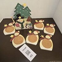 Six snowman-shaped wooden coasters with cork centers arranged around the matching snowman and Christmas tree themed holder on a dark surface.